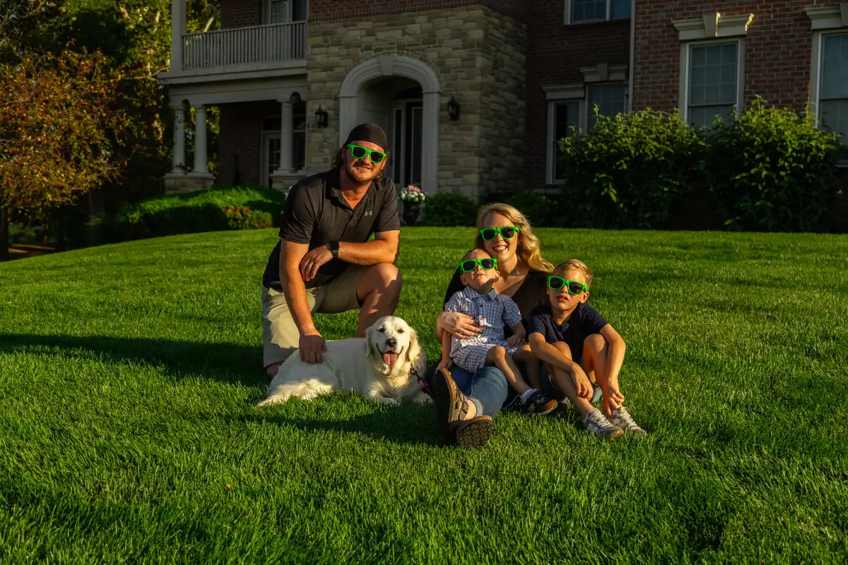 Family in green novelty glasses sitting with their white retriever on a thick front lawn