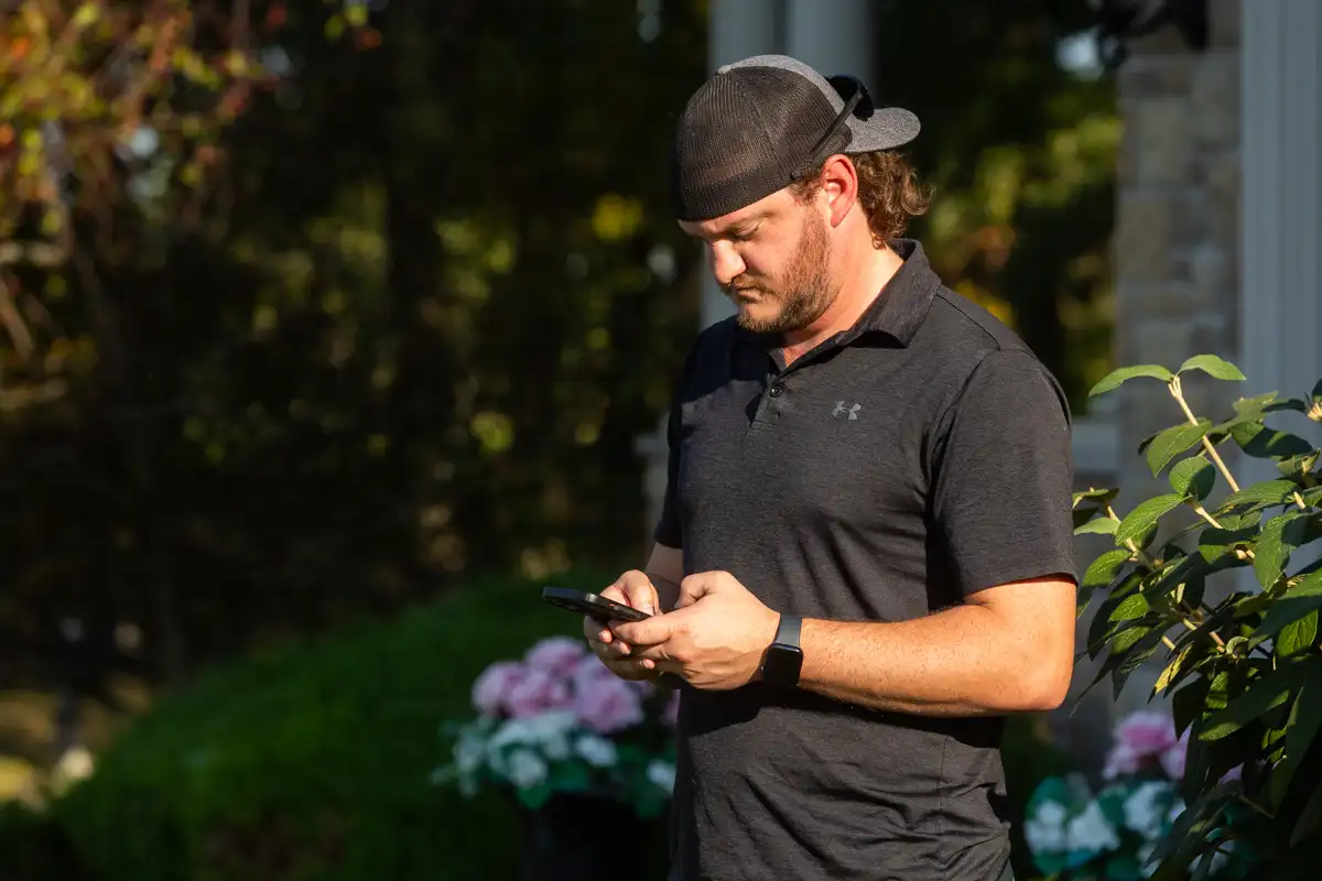 Man in a backwards cap checking his phone beside a flower bed