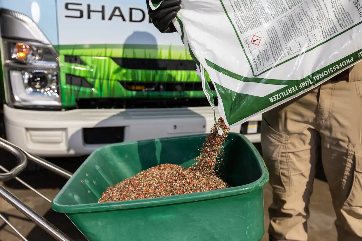 Granular fertilizer pouring from a bag into a walk-behind spreader, Shades of Green truck behind
