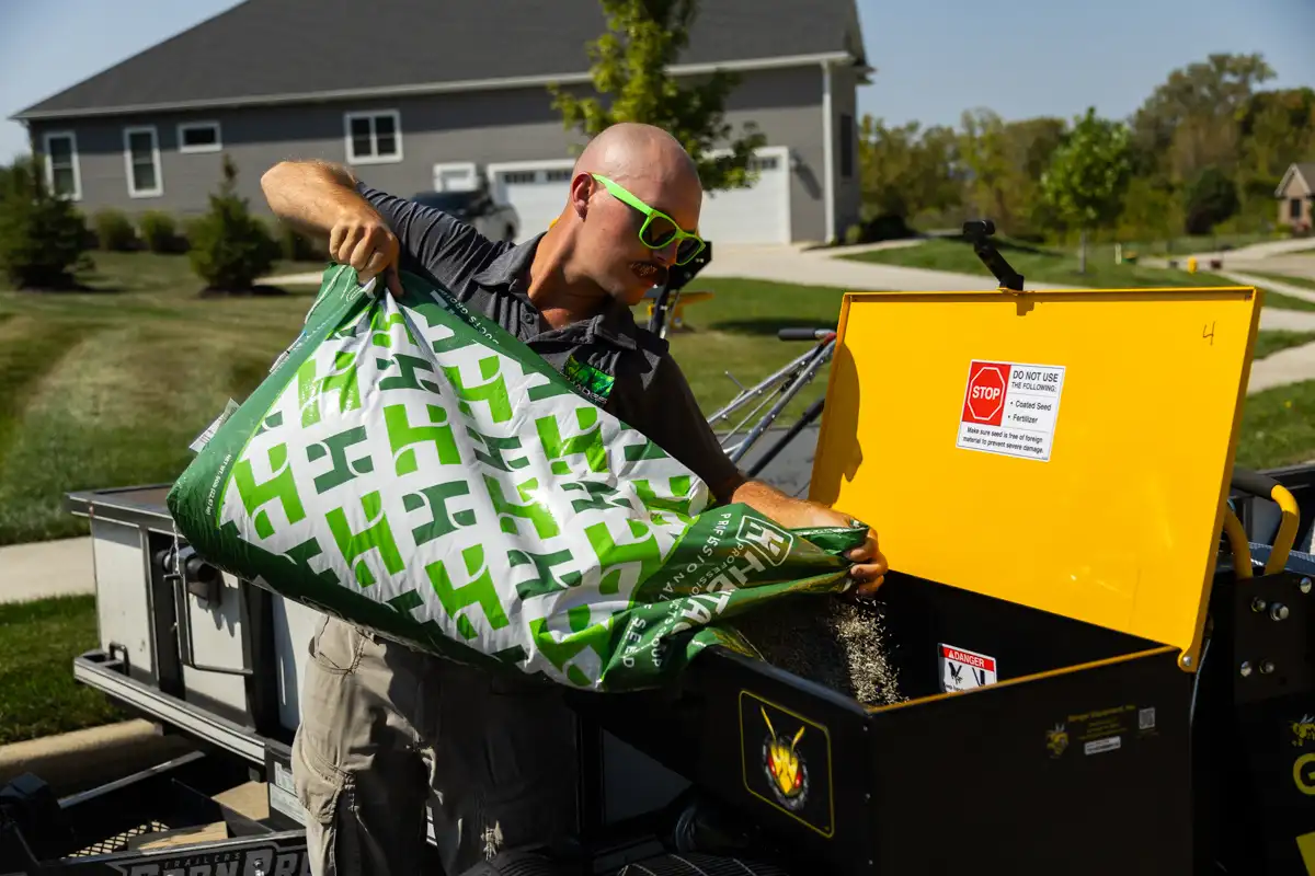 Technician in green sunglasses pouring a bag of grass seed into a spreader hopper