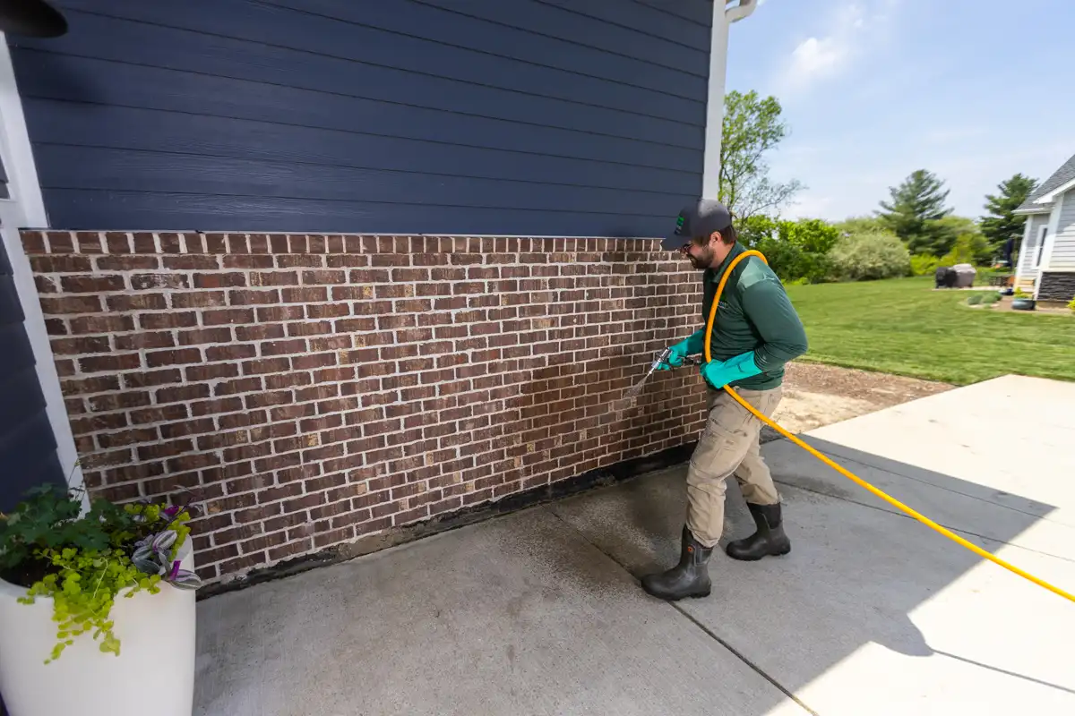 Technician spraying a perimeter pest treatment along a home's brick foundation
