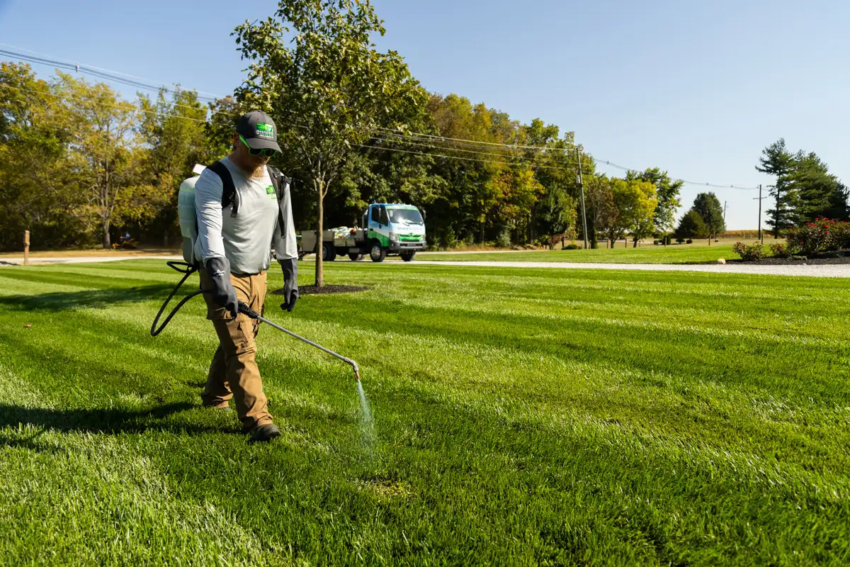 Technician with a backpack sprayer spot-treating a striped lawn, service truck behind