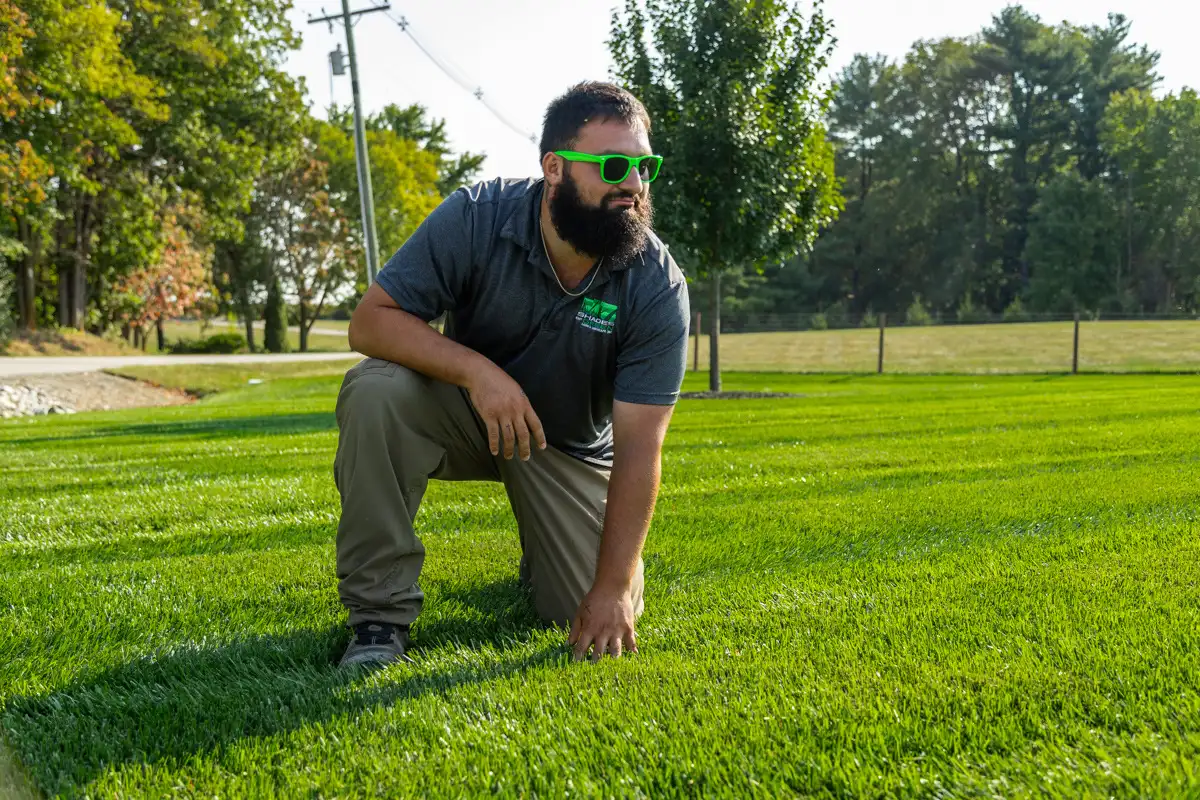 Technician in green sunglasses kneeling on a thick, striped lawn, looking across it