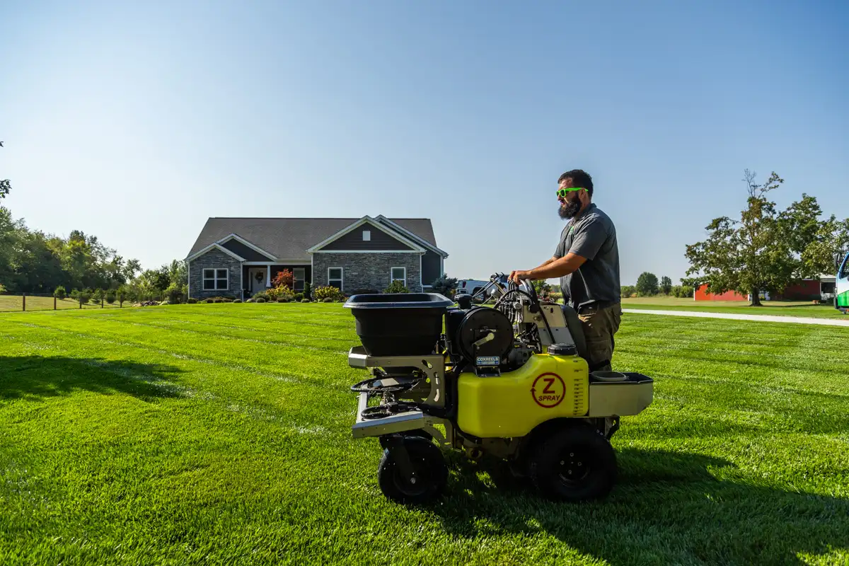 Technician on a ride-on spreader treating a wide, green lawn in front of a gray home