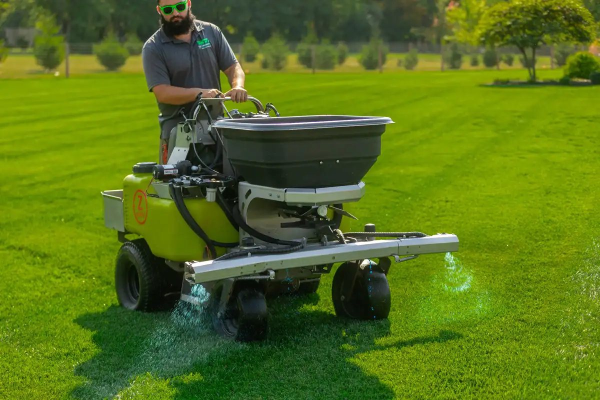 Ride-on machine's boom spraying a blue-tinted liquid treatment on a striped lawn